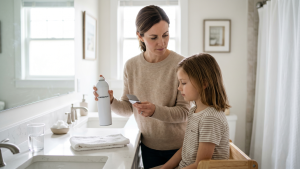 A parent stands at a bright bathroom counter holding an unbranded aerosol hairspray can and a fine-toothed metal nit comb beside her young child, quietly weighing whether to spray the can on the child's head as a lice treatment
