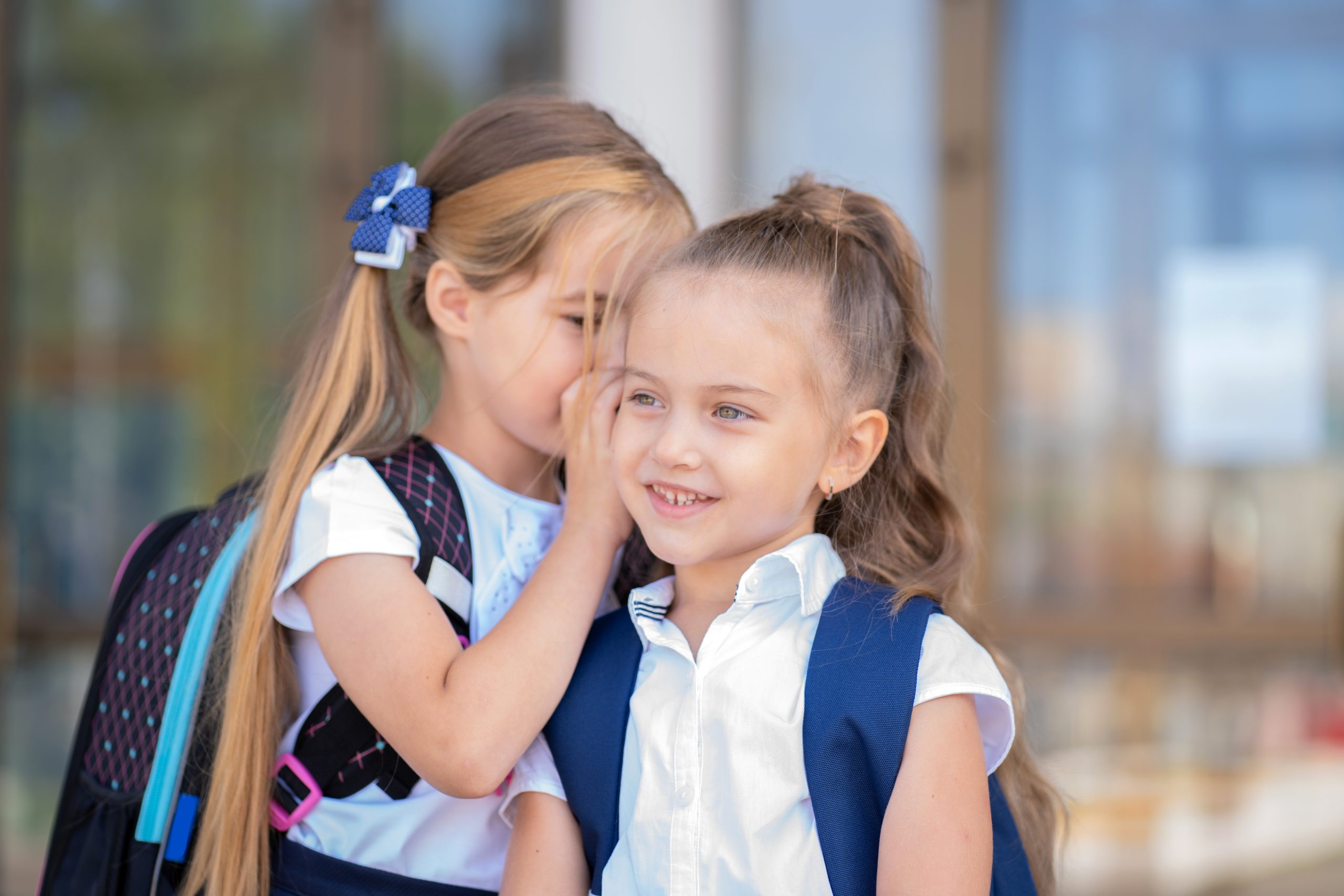 Two elementary school girls standing close together outside school while one whispers to the other, illustrating telling school about lice discreetly