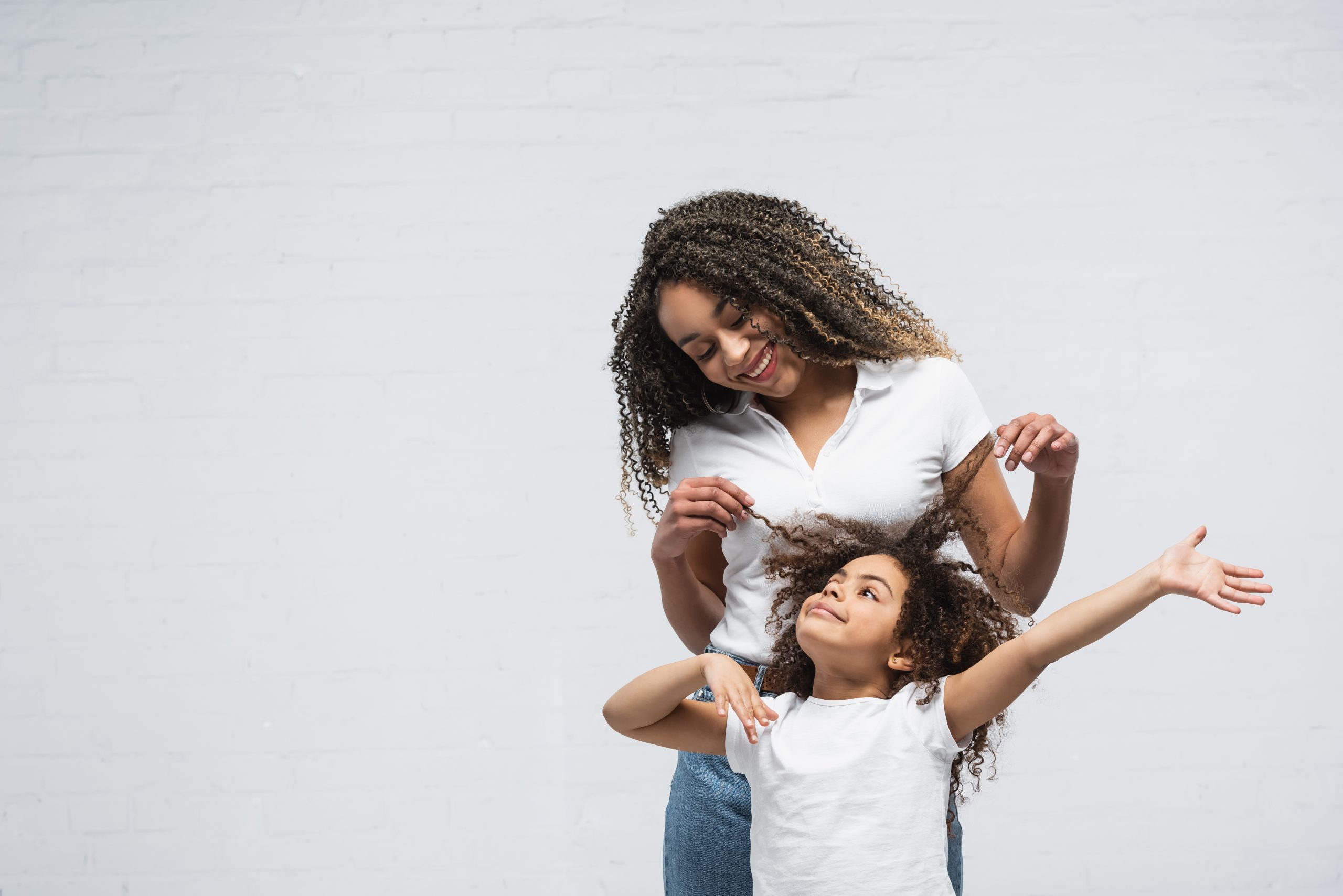 Person lifting another person's curly hair beside a white brick wall